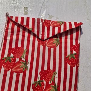 May include: A fabric bag featuring a red and white striped pattern, adorned with illustrations of strawberries. The strawberries are depicted in red with green leaves. The bag is hanging against a white wall.