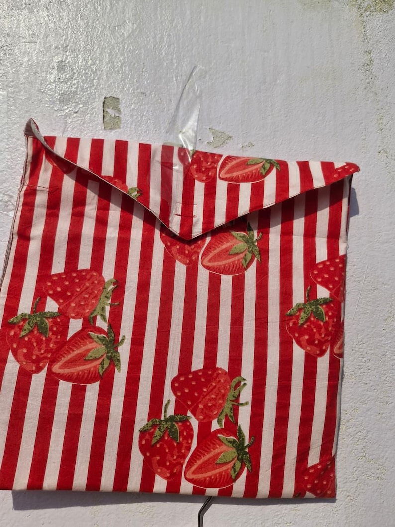 May include: A fabric bag featuring a red and white striped pattern, adorned with illustrations of strawberries. The strawberries are depicted in red with green leaves. The bag is hanging against a white wall.