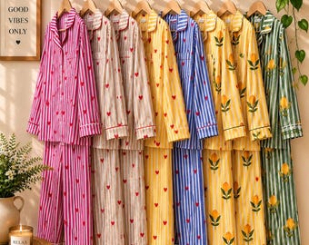 Op de afbeelding: Een collectie van acht pyjamasets met lange mouwen en knoopsluiting, hangend aan houten kleerhangers. De sets hebben diverse verticale strepenpatronen in roze, beige, geel, blauw en groen, versierd met kleine rode hartjes of gele citroenen.