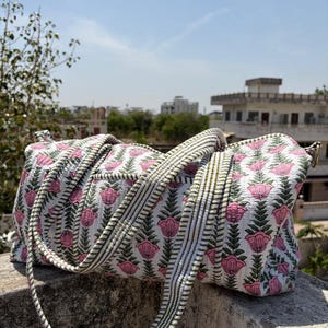 Puede incluir: Bolso de viaje blanco con estampado floral rosa y verde. El bolso presenta un patrón repetido de flores rosas y hojas verdes sobre un fondo blanco. La correa tiene rayas verdes y blancas.