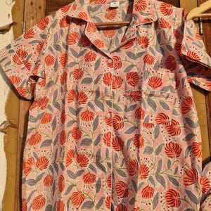 Pu&ograve; includere: Camicia da pigiama rosa a maniche corte con motivo floreale. La camicia presenta un motivo di fiori rossi e foglie grigie. Ha una chiusura con bottoni, una tasca sul petto e bordi bianchi.