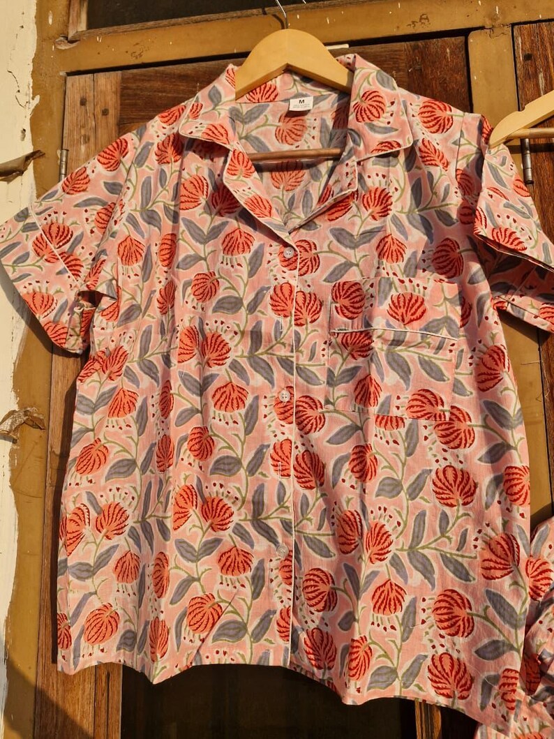 Pu&ograve; includere: Camicia da pigiama rosa a maniche corte con motivo floreale. La camicia presenta un motivo di fiori rossi e foglie grigie. Ha una chiusura con bottoni, una tasca sul petto e bordi bianchi.