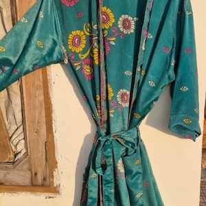 Puede incluir: Bata de color turquesa con un estampado floral en rosa, amarillo y blanco. La bata tiene mangas largas y un cinturón anudado. El diseño floral se repite en toda la prenda, creando un aspecto cohesivo y decorativo.