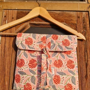 Pu&ograve; includere: Una pochette rosa con stampa floreale e chiusura con fiocco abbinata, appesa a una gruccia di legno. La pochette presenta un motivo ripetuto di fiori rossi e foglie grigie su sfondo rosa chiaro. La pochette &egrave; in tessuto.