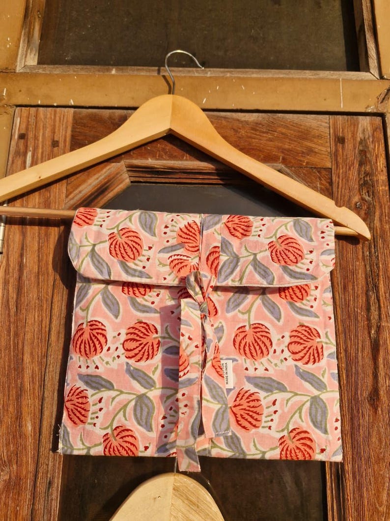 Pu&ograve; includere: Una pochette rosa con stampa floreale e chiusura con fiocco abbinata, appesa a una gruccia di legno. La pochette presenta un motivo ripetuto di fiori rossi e foglie grigie su sfondo rosa chiaro. La pochette &egrave; in tessuto.