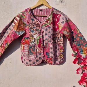 May include: A patchwork jacket in shades of pink, red, and floral patterns. The jacket features a V-neck, long sleeves, and two front pockets. It is buttoned up with small, round buttons and hangs on a wooden hanger.