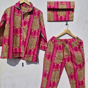 May include: A pink pajama set with a tiger print, including a button-down shirt, pants, and a matching pouch. The set features vertical stripes and a white trim. The tiger pattern is in shades of brown and tan.