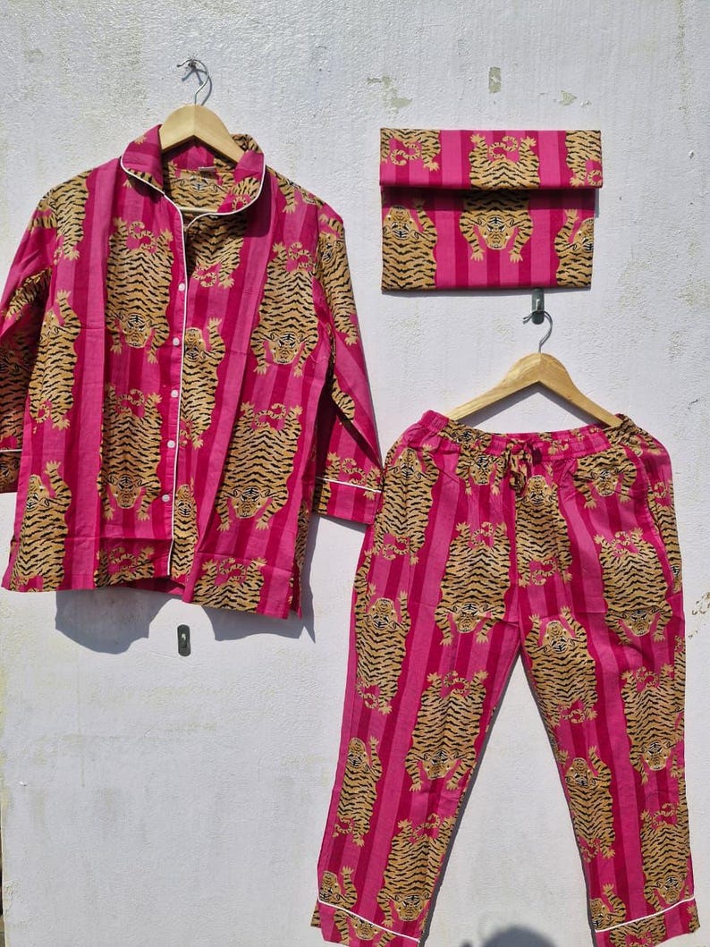 May include: A pink pajama set with a tiger print, including a button-down shirt, pants, and a matching pouch. The set features vertical stripes and a white trim. The tiger pattern is in shades of brown and tan.