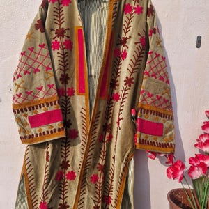 May include: A long, open-front coat in a muted olive green hue, adorned with intricate floral embroidery in shades of pink and brown. The coat features a striped lining and is trimmed with gold-colored accents. The sleeves and front panels showcase detailed stitching.
