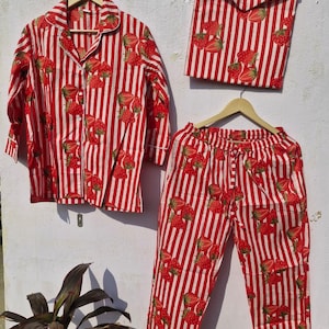 May include: A red and white striped pyjama set featuring a strawberry design. The set comprises a long-sleeved button-down shirt, trousers, and a matching pouch. The pyjamas are displayed on a wall.