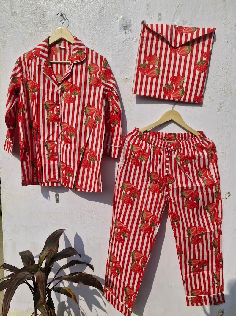 May include: A red and white striped pyjama set featuring a strawberry design. The set comprises a long-sleeved button-down shirt, trousers, and a matching pouch. The pyjamas are displayed on a wall.