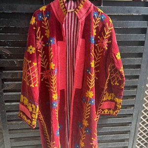 May include: A long, open-front burgundy coat with yellow and blue floral embroidery. The coat has a striped interior and is displayed on a wooden hanger. The embroidery is detailed on the collar, sleeves, and front panels. The coat is a vibrant piece.