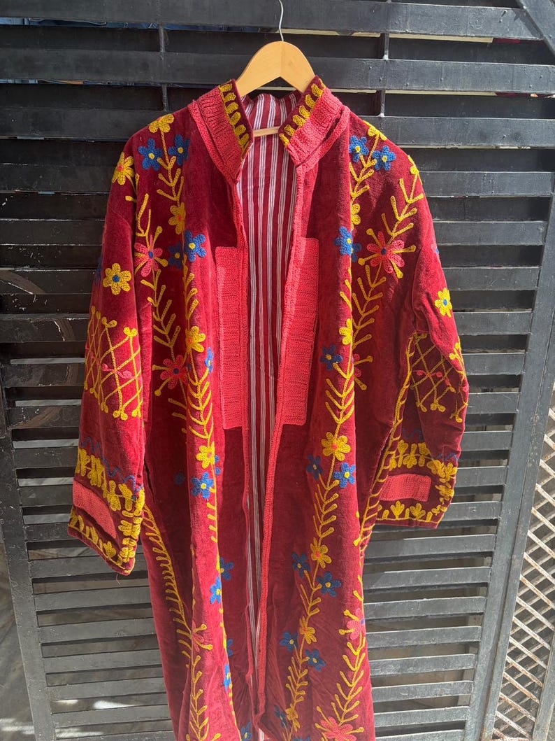 May include: A long, open-front burgundy coat with yellow and blue floral embroidery. The coat has a striped interior and is displayed on a wooden hanger. The embroidery is detailed on the collar, sleeves, and front panels. The coat is a vibrant piece.