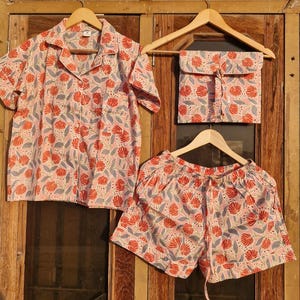 May include: A three-piece pajama set with a floral pattern. The set includes a short-sleeved button-down shirt, shorts, and a matching pouch. The fabric features a pink background with red and blue floral designs. All pieces are hanging on wooden hangers.
