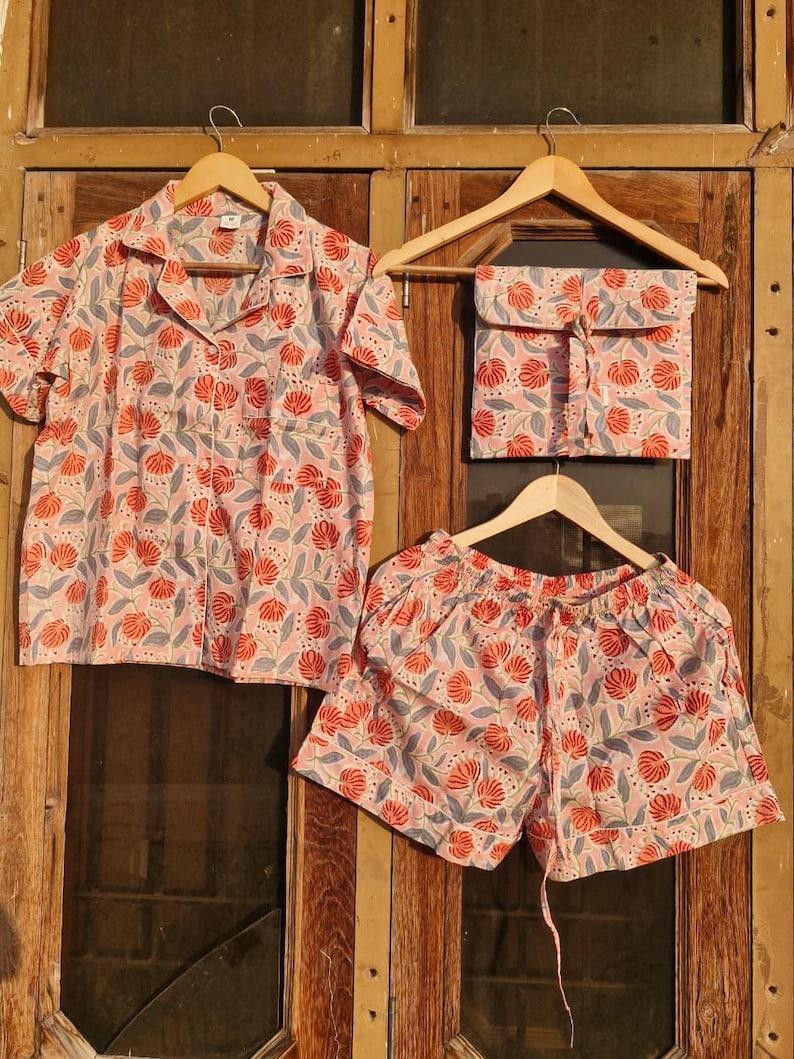 Pu&ograve; includere: Un completo pigiama a tre pezzi con motivo floreale. Il set include una camicia a maniche corte con bottoni, pantaloncini e una pochette abbinata. Il tessuto presenta uno sfondo rosa con disegni floreali rossi e blu. Tutti i pezzi sono appesi a grucce di legno.