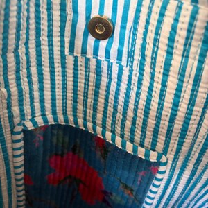 May include: Close-up of a fabric bag interior. The bag features a blue and white striped lining with a small pocket. The pocket is made of blue fabric with a floral pattern in red and pink. A gold-colored magnetic snap closure is visible.