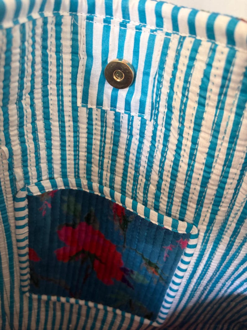 May include: Close-up of a fabric bag interior. The bag features a blue and white striped lining with a small pocket. The pocket is made of blue fabric with a floral pattern in red and pink. A gold-colored magnetic snap closure is visible.