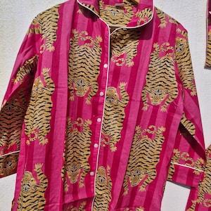 May include: A pink pajama set with a repeating tiger print. The long-sleeved top has a button-down front, a collar, and white trim. The tiger pattern is in shades of tan and brown, set against vertical pink stripes.