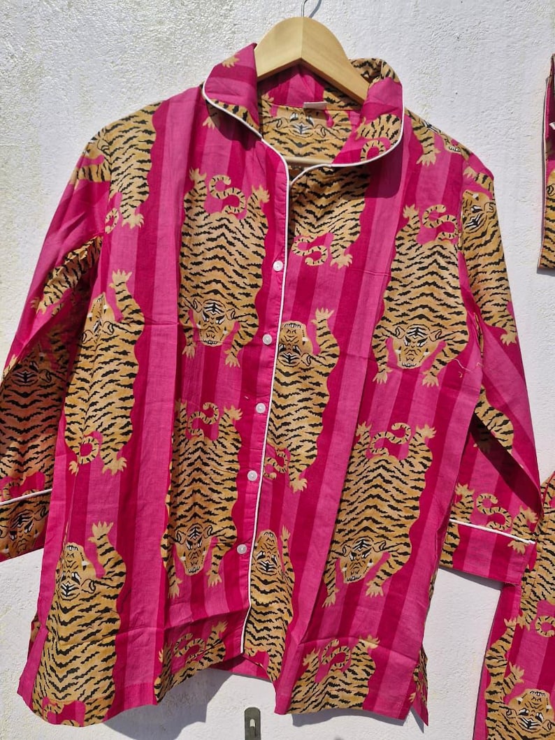 May include: A pink pajama set with a repeating tiger print. The long-sleeved top has a button-down front, a collar, and white trim. The tiger pattern is in shades of tan and brown, set against vertical pink stripes.