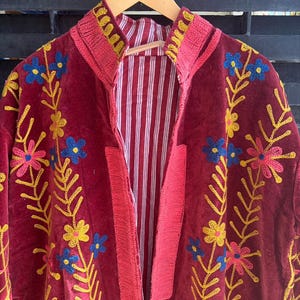 May include: A burgundy jacket with floral embroidery in yellow, blue, and pink. The jacket has a striped red and white lining and a high collar. The embroidery is detailed and covers the front and sleeves of the jacket.