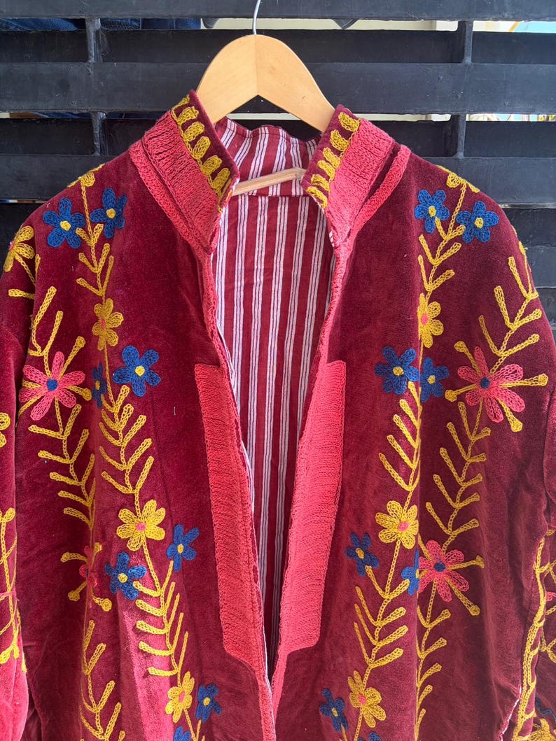 May include: A burgundy jacket with floral embroidery in yellow, blue, and pink. The jacket has a striped red and white lining and a high collar. The embroidery is detailed and covers the front and sleeves of the jacket.