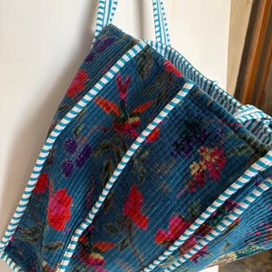 May include: A quilted tote bag with a teal background and a floral pattern in shades of red, pink, and purple. The bag has teal and white striped handles and trim. The bag is made of a soft, textured fabric.