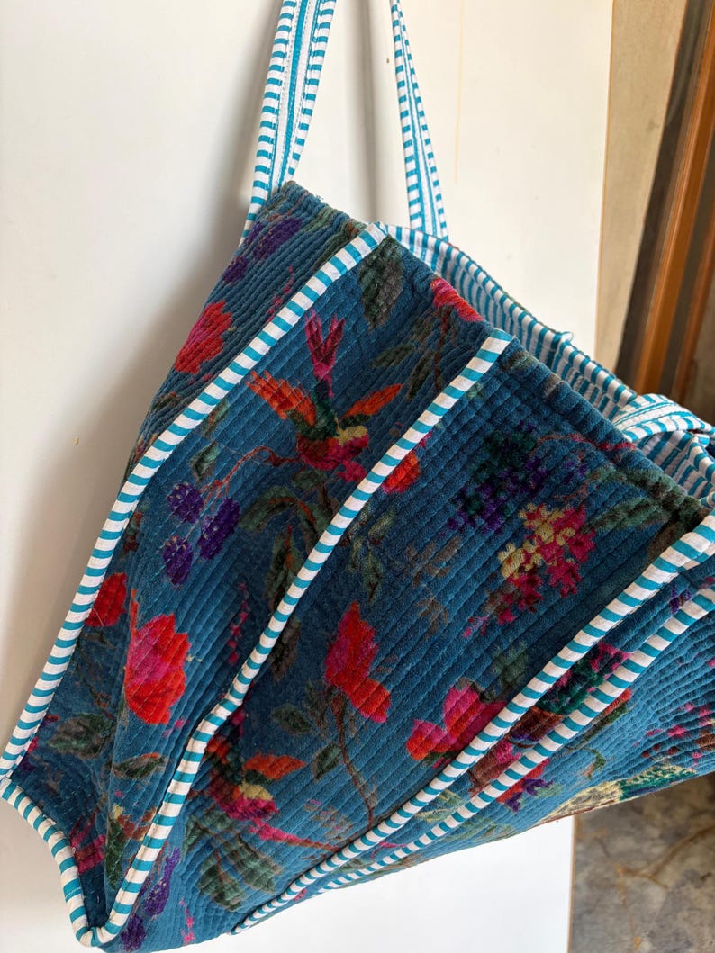 May include: A quilted tote bag with a teal background and a floral pattern in shades of red, pink, and purple. The bag has teal and white striped handles and trim. The bag is made of a soft, textured fabric.
