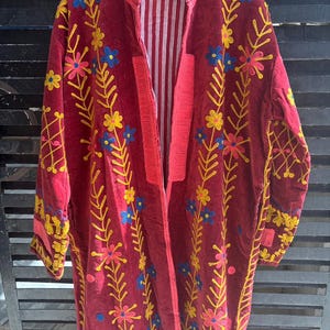 May include: A long, burgundy velvet coat with floral embroidery in yellow, pink, and blue. The coat has a striped lining and a red collar and trim. The embroidery features stylized flowers and leaves.