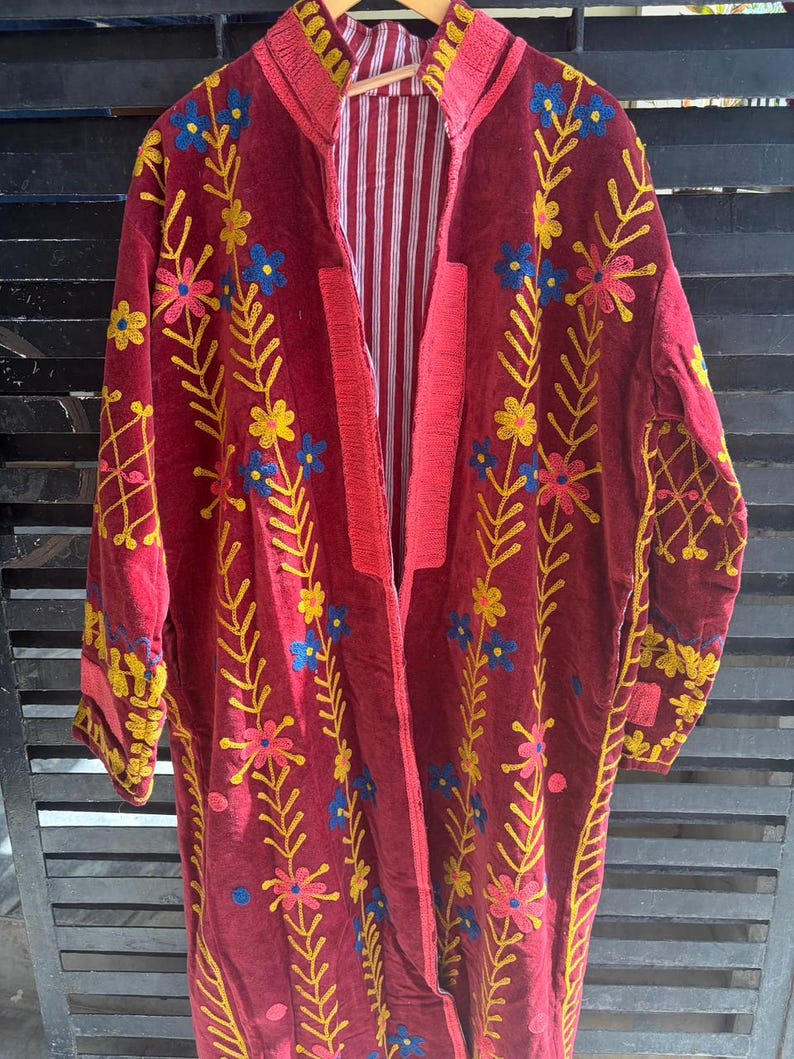 May include: A long, burgundy velvet coat with floral embroidery in yellow, pink, and blue. The coat has a striped lining and a red collar and trim. The embroidery features stylized flowers and leaves.