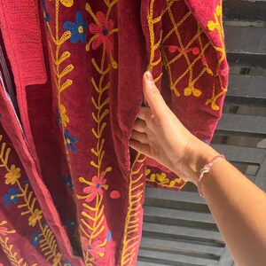May include: A burgundy robe with detailed floral embroidery in yellow, pink, and blue. The robe has a long, flowing design with detailed stitching along the edges and sleeves. The embroidery creates a vibrant contrast against the dark fabric.
