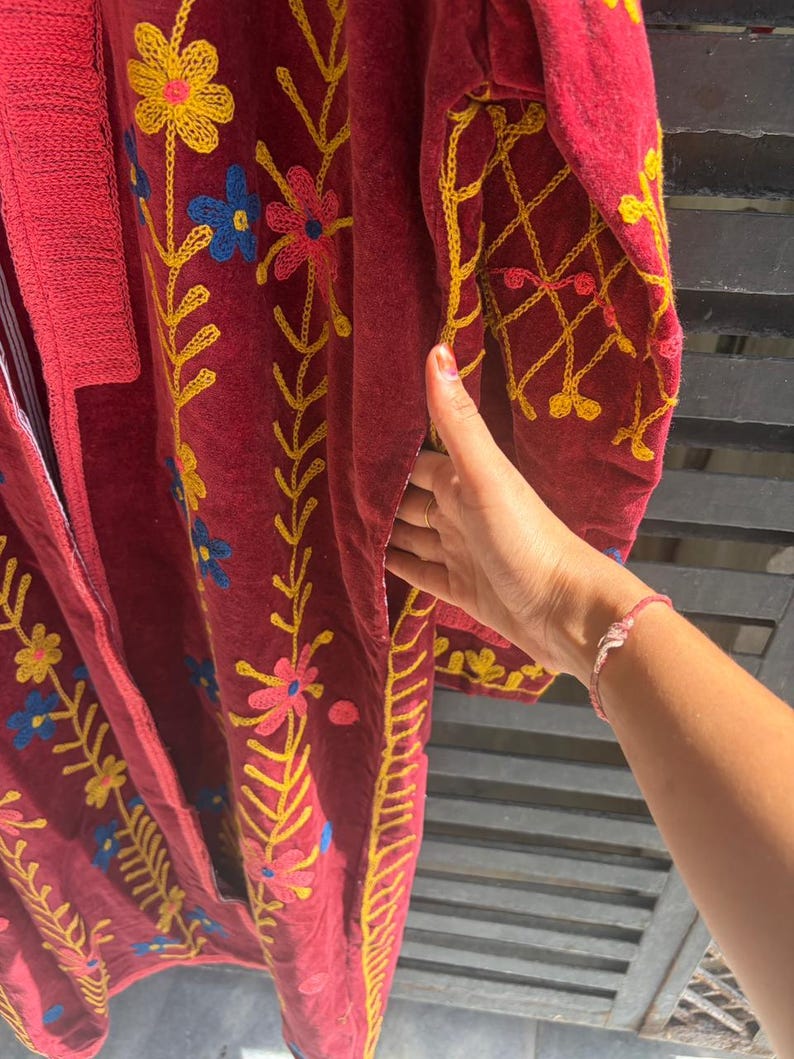 May include: A burgundy robe with detailed floral embroidery in yellow, pink, and blue. The robe has a long, flowing design with detailed stitching along the edges and sleeves. The embroidery creates a vibrant contrast against the dark fabric.