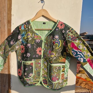 May include: A quilted jacket with a patchwork design featuring floral and paisley patterns in shades of green, pink, and blue. The jacket has a green striped lining, two front pockets, and a button closure. The jacket is hanging on a wooden hanger.