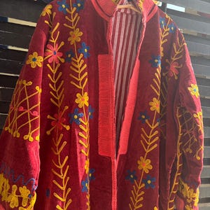 May include: A burgundy embroidered jacket with floral patterns in yellow, blue, and pink. The jacket has a red trim and a striped white and red lining. The jacket is hanging on a wooden hanger.