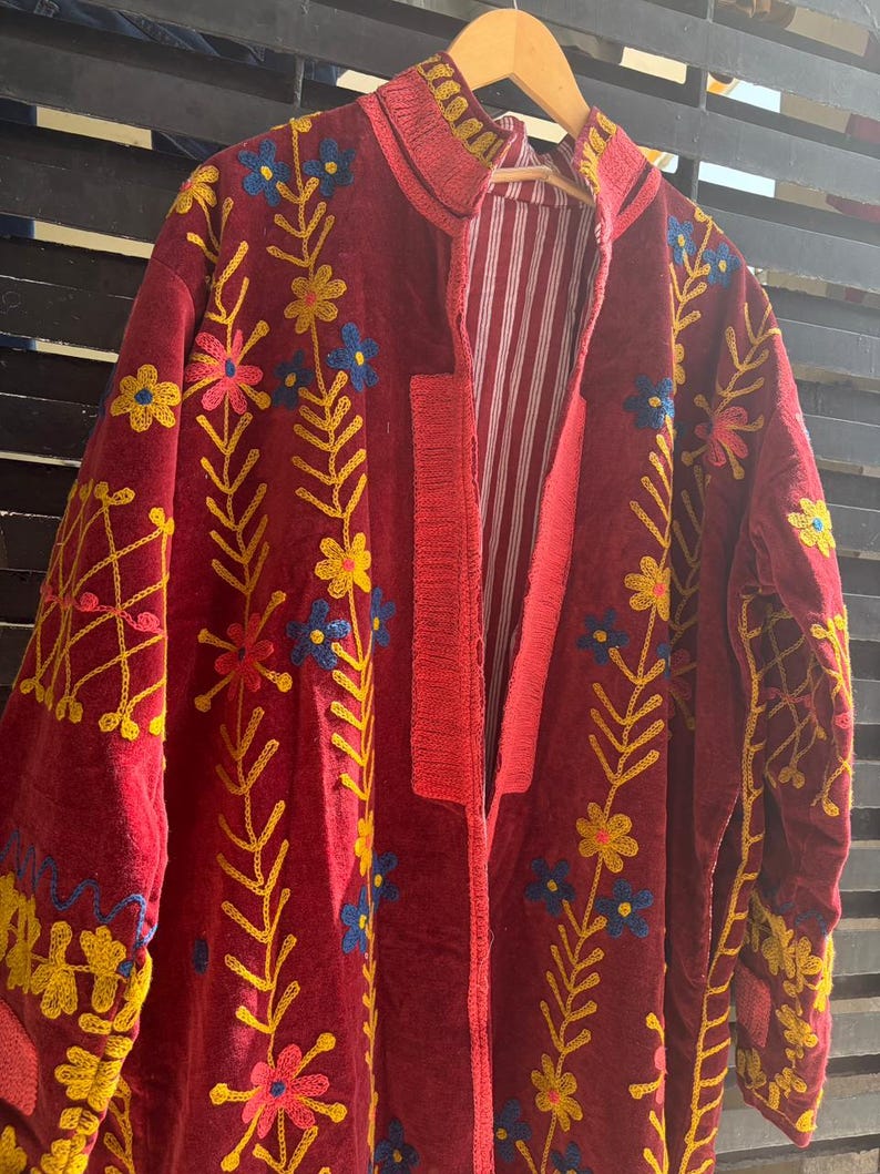 May include: A burgundy embroidered jacket with floral patterns in yellow, blue, and pink. The jacket has a red trim and a striped white and red lining. The jacket is hanging on a wooden hanger.