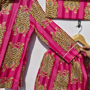 May include: Pink pajama set with a tiger print. The set includes a long-sleeved top, pants, and a folded item, all featuring a repeating pattern of tigers on pink vertical stripes. The top and pants have white trim.