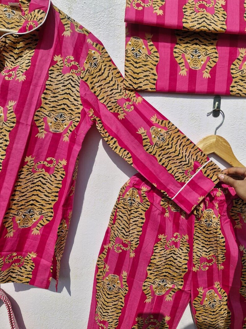 May include: Pink pajama set with a tiger print. The set includes a long-sleeved top, pants, and a folded item, all featuring a repeating pattern of tigers on pink vertical stripes. The top and pants have white trim.