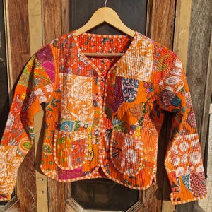 Op de afbeelding: Een oranje patchwork jasje met een V-hals en lange mouwen. De jas heeft een verscheidenheid aan kleurrijke bloemen- en geometrische patronen. Het heeft een knoopsluiting en twee afgeronde zakken. Het jasje hangt aan een houten hanger.