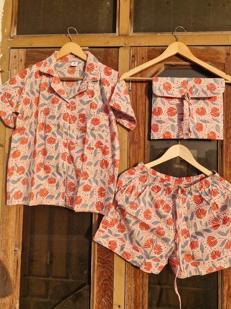 Pu&ograve; includere: Un completo pigiama con fantasia floreale. Il set comprende una camicia a maniche corte, pantaloncini e un piccolo astuccio. Il tessuto &egrave; rosa chiaro con un motivo ripetuto di fiori rossi e foglie grigie. Appeso a grucce di legno.