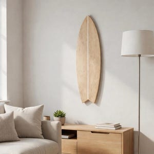 May include: A wooden surfboard with a light brown finish and a white stripe down the center is mounted on a white wall. The surfboard is displayed vertically in a living room setting, with a sofa, lamp, and wooden cabinet.