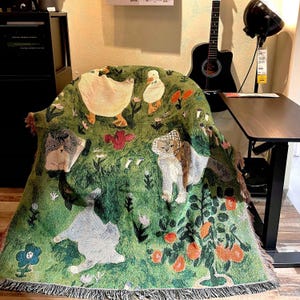 May include: Green throw blanket with a whimsical design featuring cats, ducks, flowers, and fruit trees. The blanket has a fringed edge and is draped over a chair. The color palette includes shades of green, white, orange, and brown.