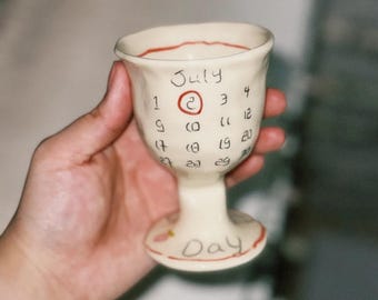 May include: A handmade ceramic goblet with a calendar design for July. The goblet is off-white with red accents and features the word "July" and the date "2" circled in red. The word "Day" is written on the base.