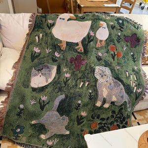 May include: Green throw blanket with a whimsical design featuring ducks, cats, and floral elements. The blanket has a fringed edge and is draped over a white sofa. The design includes various flowers and plants.