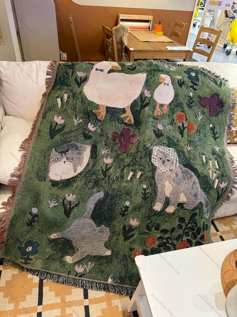 May include: Green throw blanket with a whimsical design featuring ducks, cats, and floral elements. The blanket has a fringed edge and is draped over a white sofa. The design includes various flowers and plants.