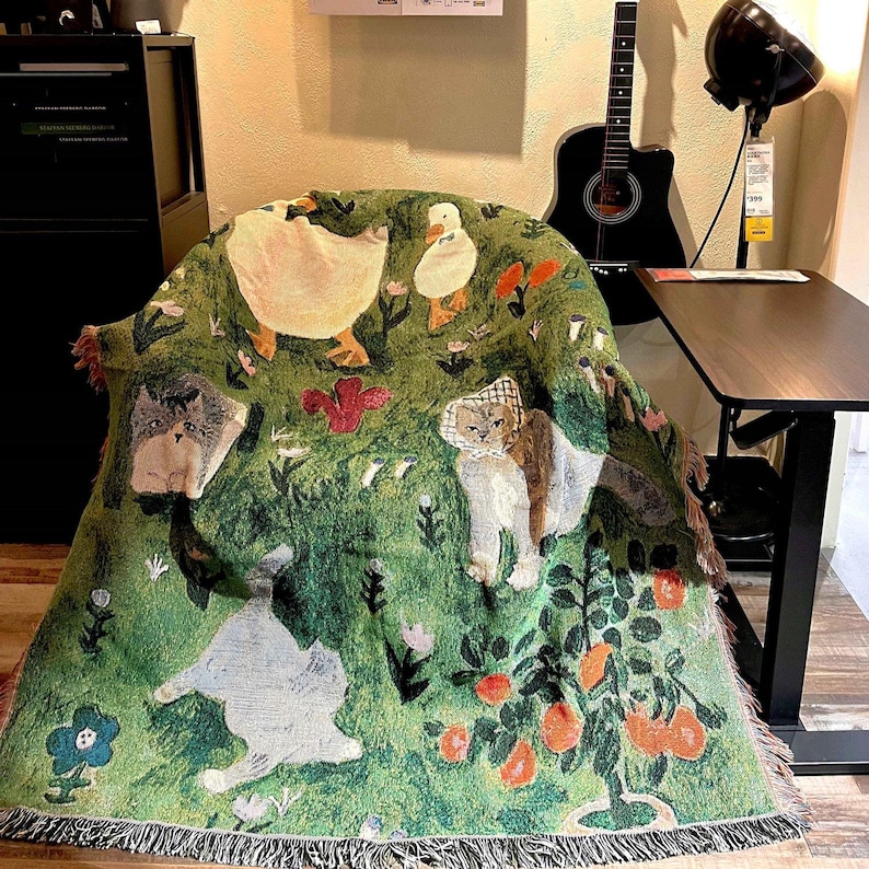 May include: Green throw blanket with a whimsical design featuring cats, ducks, flowers, and fruit trees. The blanket has a fringed edge and is draped over a chair. The color palette includes shades of green, white, orange, and brown.