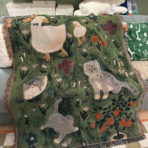 May include: Green throw blanket featuring a whimsical design of geese, cats, flowers, and a small potted tree. The blanket has a fringed edge and is made of a soft, woven material. The design includes a large goose wearing a bow tie.