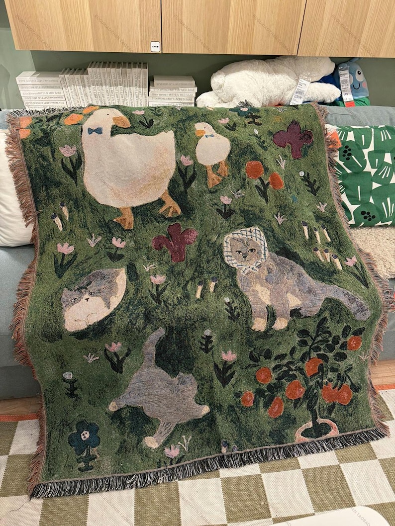 May include: Green throw blanket featuring a whimsical design of geese, cats, flowers, and a small potted tree. The blanket has a fringed edge and is made of a soft, woven material. The design includes a large goose wearing a bow tie.