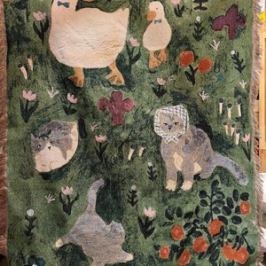 May include: Green woven throw blanket featuring whimsical illustrations of ducks, cats, flowers, and plants. The blanket has a fringed edge and a textured appearance. The color palette includes shades of green, cream, orange, and purple.