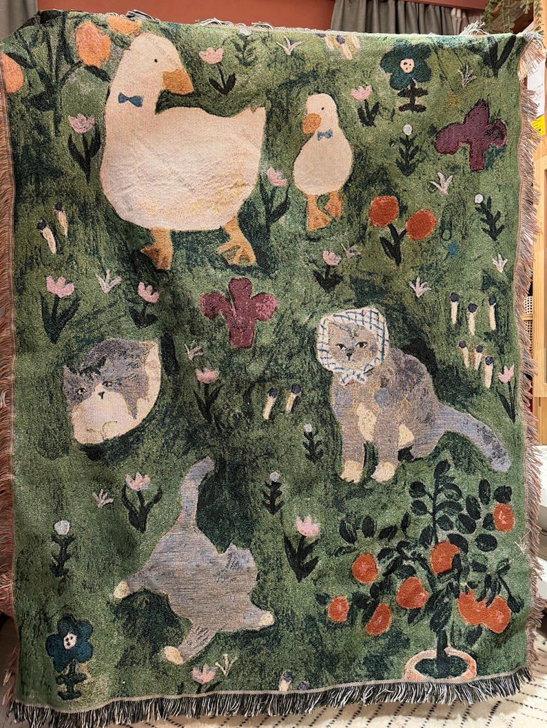 May include: Green woven throw blanket featuring whimsical illustrations of ducks, cats, flowers, and plants. The blanket has a fringed edge and a textured appearance. The color palette includes shades of green, cream, orange, and purple.