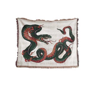 May include: Off-white throw blanket featuring a detailed illustration of two intertwined snakes. The snakes are primarily green and red, with intricate scales and a menacing expression. The blanket has a fringed edge.