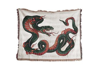 May include: Off-white throw blanket featuring a detailed illustration of two intertwined snakes. The snakes are primarily green and red, with intricate scales and a menacing expression. The blanket has a fringed edge.